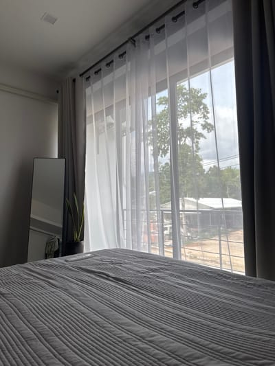 The image shows an interior view of a room, likely a bedroom, in Moo 4, Thailand, as can be inferred from the outdoor scenery visible through a window. The foreground contains a portion of a bed covered with a gray, vertically striped comforter. A window runs horizontally across the upper portion of the frame, covered by translucent white curtains attached to a dark rod with visible rings. The outside view includes trees, buildings, and electrical wires, typical for an urban or suburban setting in Thailand. A mirror, mounted on the wall, reflects a plant and a storage area. The walls are painted white. The overall scene suggests a simple, modern bedroom with a view of the surrounding Moo 4 area.