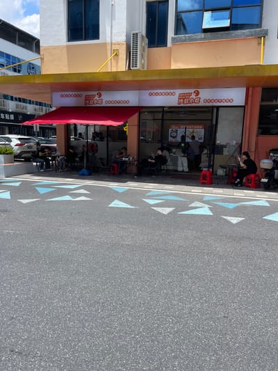 In Panyu District, China, a bustling street scene unfolds in front of a restaurant named "SHAXIAN SNACKS". The restaurant, adorned with a vibrant red awning, is open to the street, with patrons seated at tables both inside and outside. Several individuals are seen eating and interacting, with one person using a mobile phone. A red stool is placed outside the entrance, and a motorcycle is parked nearby. The pavement is decorated with blue and white triangular patterns. The building facade features large windows and an air conditioning unit. The sky above is a bright blue with scattered clouds, suggesting a clear day.