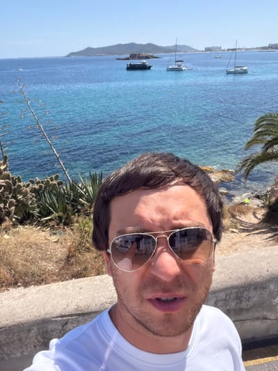 A man wearing aviator sunglasses and a white t-shirt takes a selfie outdoors with the sea and coastline in the background. The sea is a vibrant turquoise and blue, dotted with several sailboats. A dark-hulled boat is anchored closer to the shore, and a distant coastline with buildings is visible. The foreground features dry brush, cacti, and a concrete barrier. The man's sunglasses reflect a white building and a car. The scene appears to be in Eivissa, Spain, given the sunny, coastal environment and typical Mediterranean vegetation.