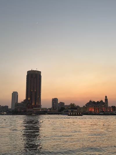 A dusk view of the Cairo skyline features the Nile River in the foreground, with a crescent moon visible in the pale, gradient sky. On the riverbank, a tall, cylindrical hotel building with the "SOFITEL" logo is illuminated, as are several other buildings and a mosque with a minaret. The water reflects the ambient light of the sky and the city, and a few small objects, possibly debris or small boats, are visible on its surface. The scene is likely captured from a boat on the Nile, looking towards the city's eastern bank.