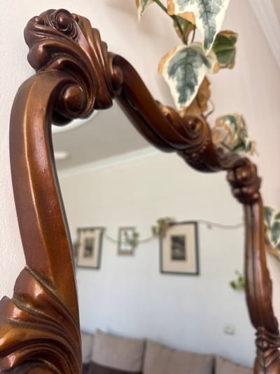 The image is a close-up, eye-level, medium shot of a decorative wooden mirror frame with an ornate carved design, set against a plain white wall. A variegated ivy plant with green and white leaves drapes over the top and right side of the mirror. The mirror reflects a room with a white wall, two framed pictures, and the back of a brown sofa. The overall aesthetic suggests an indoor setting, likely a home. There is no indication of the city of Gyumri, Armenia, or any specific location within it.