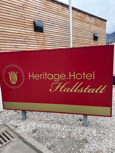 An outdoor sign for "Heritage.Hotel Hallstatt" is prominently displayed in the foreground, indicating its location in Hallstatt, Austria. The sign is a rectangular red panel with gold lettering and a gold horizontal stripe at the bottom. A circular gold emblem, resembling a stylized "W" or tulip, is positioned on the left side. The sign is supported by two grey metal posts embedded in a bed of white gravel. To the lower left, a concrete curb and a metal storm drain grate are visible. In the background, a building with a facade constructed from horizontal wooden slats extends across the upper portion of the frame. Two black, rectangular vent covers are mounted on this wooden facade. A thin, vertical white flagpole stands between the sign and the building. Distant mountains are faintly visible under an overcast sky in the upper right quadrant. No individuals or active subjects are present in the scene.