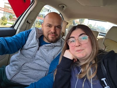 The image displays two individuals inside a vehicle in Gevgelija, North Macedonia. A bald, bearded male, wearing a blue long-sleeved shirt and a grey quilted vest, is seated on the left, looking directly at the camera with a slight smile. To his right, a female with long, light brown hair and round eyeglasses is seated, wearing a dark hooded sweatshirt under a black zippered jacket. She rests her chin on her left hand and looks towards the camera with a neutral expression. The vehicle interior features light-colored upholstery on the seats and ceiling. Through the windows, a bright sky, green foliage, distant buildings, and bare trees are visible. A red object is partially visible through the upper portion of the driver's side window.