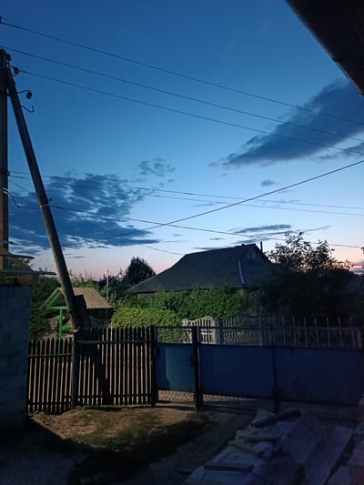 The image depicts a rural twilight scene in Leușeni, Moldova, characterized by a predominantly blue sky with scattered clouds. A prominent wooden utility pole with overhead wires stands to the left of the frame, leaning slightly. In the background, a dark-roofed house with a visible chimney is situated behind a dense green hedge. A wooden fence with vertical slats separates the foreground from the background, and a wider, dark blue gate is visible to the right of the wooden fence. The lighting suggests it is either dusk or dawn.