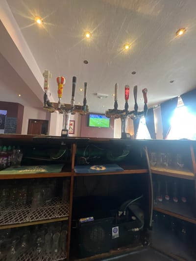 A bar features multiple beer taps, including Foster's and Amstel, mounted on a wall with recessed lighting. A TV screen displays a game in the background. Behind the taps, a dark wooden cabinet houses beer equipment, including a cooling unit with visible tubes and wires. A glass rack filled with glasses sits on a lower shelf of the cabinet, along with beer branded display pieces.
