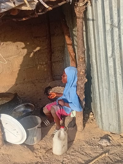 A young person in a light blue head covering and shirt, and pink shorts, is squatting inside a rustic structure with a corrugated metal roof supported by wooden beams. Next to them, there is a white plastic jug, black and white striped sandals, and a stack of bowls and a lid. The floor is dirt, and the walls appear to be made of mud brick. The lighting is dim, highlighting the interior's simple and possibly impoverished conditions.