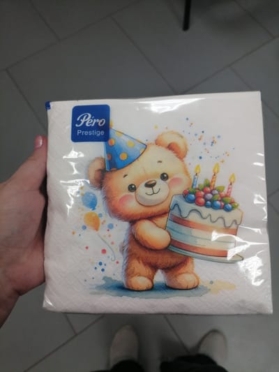 A hand with a pink fingernail holds a stack of white paper napkins featuring a watercolor illustration of a teddy bear wearing a blue party hat and holding a birthday cake. The cake is decorated with red and blue berries and has three lit candles. Colorful confetti and balloons are scattered around the bear. A blue and white logo that reads "Pero Prestige" is visible in the upper left corner of the napkins. The napkins are enclosed in clear plastic packaging. The background is a gray tiled floor and a dark gray ceiling with visible seams, suggesting an indoor environment. The scene appears to be staged, with the hand and napkins centrally positioned. The context provided regarding "Смалявічы, Belarus" does not align with the visual information in the image, as there are no discernible landmarks or clues to suggest this specific location.