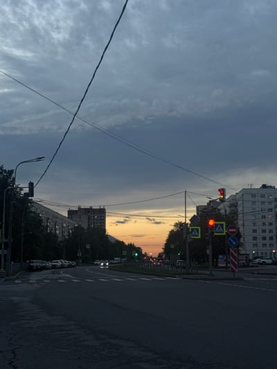 This is a medium-shot photograph of a street scene in Ladozhskoe Ozero, Russia, taken at dusk. The sky is filled with heavy, dark clouds that are beginning to break, revealing a sliver of orange and yellow sunset on the horizon. Power lines crisscross the sky. A street light with two fixtures is visible on the left, and a traffic light on the right displays a red signal. Several cars are parked along the left side of the road, and a few cars are driving on the road, their headlights illuminated. Pedestrian crossing signs are present at the intersection. Tall apartment buildings line the street, with lights visible in some windows. Trees are interspersed along the roadside. The road surface appears wet and slightly cracked.