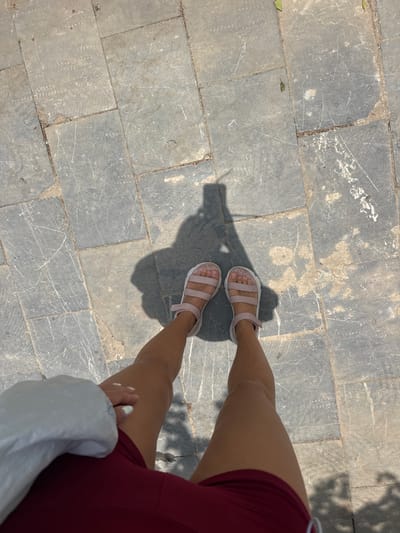 An eye-level shot shows a section of a paved area with a person standing on it. The paving consists of gray, rectangular stones with visible scratches and imperfections. The person's legs, clad in light pink sandals, are positioned in the center of the frame. They are wearing maroon shorts. A portion of a white garment, possibly a jacket or bag, is visible in the lower right corner. The location is Mèo Vạc Commune, Vietnam.
