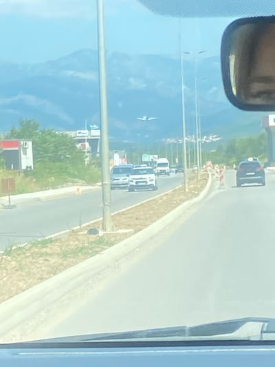 The image depicts a view from inside a vehicle traveling on a road in Radanovići, Montenegro. The road appears to be a highway with multiple lanes. On the left side of the road, there are businesses with signage and greenery. In the distance, mountains are visible under a bright sky. An airplane is seen in the sky, suggesting the proximity of an airport. Several cars and vans are on the road, indicating moderate traffic. Construction barriers are present along the side of the road. The scene appears to be captured during the daytime.