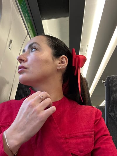 A female subject is depicted in a close-up, profile view, positioned within a modern train carriage. She has dark hair, styled into a ponytail secured with a prominent red bow. Her visible right earlobe displays three small, light-colored stud earrings. She wears a bright red garment, consistent with a jacket or blouse, featuring a visible pocket flap on the right chest area. Her left hand, adorned with a ring featuring a light-colored oval stone on the ring finger and a thin, gold-colored bracelet on the wrist, is raised to her neck/collar, gently touching the fabric. Her head is tilted slightly upwards, and her gaze is directed towards the upper left of the frame. The background reveals typical train interior elements: a window frame with a green "SOS" sign on the left, white strip lighting integrated into dark grey ceiling panels, and a dark grey, patterned seatback partially visible on the right. No specific landmarks or external features are visible to pinpoint a precise location within the city of Bath, United Kingdom; the scene is identifiable only as an interior of a train carriage.