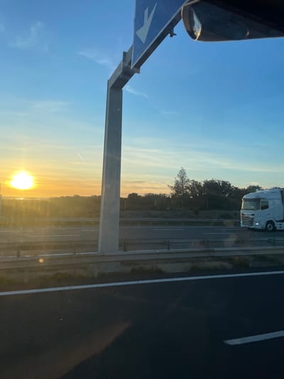 The image depicts a multi-lane highway near La Palme, France, at either sunrise or sunset. The sky displays a clear gradient, transitioning from a bright yellow-orange at the horizon, where a distinct, intensely bright sun is visible on the left, to a clear blue overhead, with a single faint aircraft contrail present.
In the foreground, a dark asphalt road surface with a white solid lane marker is visible from the perspective of a vehicle, likely in motion. A grey concrete barrier separates this immediate lane from the main highway. A vehicle's rearview mirror is partially visible in the upper right.
The midground features the multi-lane highway, bordered by metal guardrails and additional concrete barriers. A large white semi-truck, specifically an IVECO, travels in a parallel lane from right to left across the frame. A substantial grey concrete gantry structure spans the highway, with its central vertical support prominent. A blue overhead road sign, featuring a white downward-pointing arrow, is partially visible attached to this gantry. Sparse vegetation, including dry grass and low shrubs, lines the road shoulders.
In the background, a body of water, reflecting the intense sunlight, is visible on the far left horizon. Denser, dark green trees and scrubland extend along the right side of the highway.