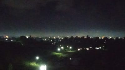 This media file depicts a nighttime landscape with a distant cityscape. The scene is dark, suggesting it is late at night or early in the morning, with a hazy, light-polluted sky visible above the horizon. The foreground is dominated by dark silhouettes of trees and undulating terrain, with several scattered artificial lights illuminating patches of ground, possibly indicating pathways or individual dwellings.

In the mid-ground, a more concentrated collection of lights suggests a settlement or town. These lights vary in color and intensity, with some appearing yellowish and others white, hinting at different types of illumination or buildings. The silhouette of trees is also prominent in this area, interspersed with the lights. The distant horizon is marked by a continuous band of light, characteristic of a larger urban or semi-urban area. A faint glow is present in the sky, contributing to the overall dark and atmospheric mood of the image.

There are no visible people or specific objects that stand out, making the scene feel vast and impersonal. The weather appears to be clear enough to allow visibility of the distant lights, though the haze in the sky might suggest some atmospheric moisture or pollution. The overall impression is one of a quiet, nocturnal view of a populated area from a slightly elevated vantage point, likely a rural or semi-rural location overlooking a town or city. The mood is serene and perhaps a little mysterious due to the darkness and the indistinct nature of the scene. No text is visible in the image.