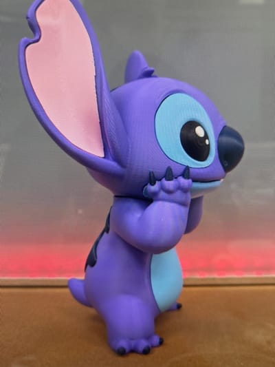 The image shows a close-up, side-view of a purple figurine, likely a character from a cartoon, placed on a brown surface. The figurine is detailed with large blue eyes, a dark nose, and pink inner ears. Its right hand is raised to its mouth, with sharp black claws visible. The background is a blurred gradient of grey at the top, transitioning to a vibrant pink and red hue at the bottom, suggesting a sunset or artificial lighting. There is no indication of the city of Kamianske, Ukraine, or any urban environment.