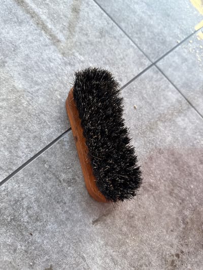 A dark-bristled brush with a wooden handle lies on a tiled surface. The tiles are rectangular and grey with dark grout lines. The brush is positioned diagonally across the frame, with its bristles facing upwards and to the right. The surface appears to be outdoors, possibly a patio or walkway, given the texture and grout lines of the tiles. There are no people visible in the image. The lighting suggests it is daytime. The image is a close-up shot, focusing on the brush and its immediate surroundings. The specific location of Białystok, Poland, cannot be determined from the visual information present in the image.