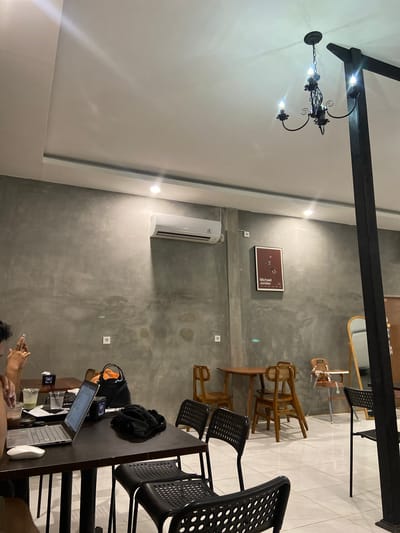 The image shows an indoor scene, likely a cafe or restaurant, with a contemporary, industrial aesthetic. The walls are a rough, textured gray concrete, and the ceiling is white. A dark wooden beam spans across the upper part of the image, from which hangs a decorative black chandelier.
The space is furnished with several wooden chairs, some with woven seats, and black perforated chairs. A long, dark wooden table is set up, with a laptop open, a computer mouse, and drinks on it. A backpack and an orange bag are also on the table. Two people are partially visible, one appearing to use a phone and another working on the laptop.
On the gray wall, a framed poster with the text "Michael Jordan" is visible. An air conditioning unit is mounted on the wall, suggesting a climate-controlled environment. The lighting appears to be artificial, with the chandelier and perhaps spotlights casting light. The overall atmosphere suggests a relaxed, perhaps studious or work-oriented environment. The location context indicates Sekar Mawar, Indonesia, but there are no specific visual cues to confirm this beyond the provided information. The time of day is not discernible.