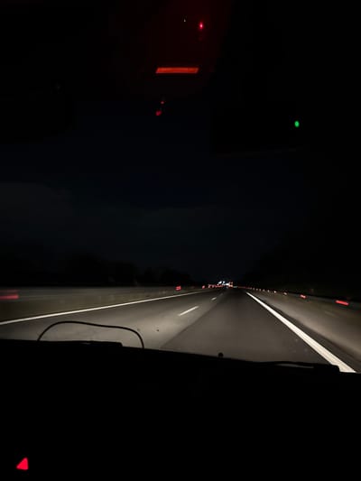 The image depicts a nighttime view from a moving vehicle, possibly a car or truck, on a highway. The right side of the image shows the highway illuminated by headlights, including white lane markings and possibly other vehicles' tail lights in the distance. The left side of the image is dark, with glimpses of what appears to be a dashboard or interior of the vehicle, including faint lights. The scene suggests nighttime travel on a well-lit highway, but lacks specific identifying details to determine a location in Manole, Bulgaria.