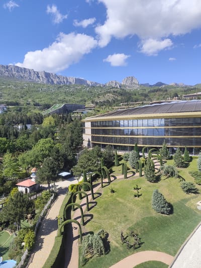 The image depicts a view in Симеизский поселковый совет, Ukraine, featuring a large modern building with reflective windows, a curved roof, and a gold-toned trim. The building is surrounded by a meticulously maintained landscape, including shaped bushes, archways formed by greenery, and winding paths. The foreground consists of various trees and shrubs, a small building with a red-tiled roof and a dome, and a fenced walkway. In the background, there are green hills and mountains under a blue sky with scattered white clouds. Another building with a sloping green roof can be seen nestled among the trees in the middle ground.