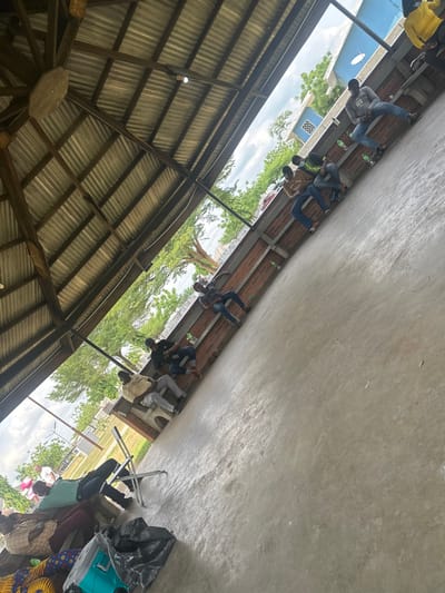 The scene depicts a large, open-sided shelter in Ozuaha, Nigeria, with a corrugated metal roof supported by wooden beams and a light-grey concrete floor. A low brick wall, serving as a bench, runs along the right side of the structure. Beyond the shelter, green trees and foliage are visible under an overcast sky, with distant buildings, including one distinctive blue structure featuring a white satellite dish.
Approximately twelve individuals, predominantly adult males, are present within the shelter. Most are seated along the brick wall or on the concrete floor. One person is lying prone on the concrete floor in the lower-left foreground, their head resting on bundled colorful fabric, with a white folding chair positioned nearby. Another individual is seated on a white plastic chair to the left. Other individuals are seated along the brick wall, some with plastic bottles placed nearby. One individual seated on the brick wall towards the right appears to be wearing a face mask. A blue hardshell suitcase and a black plastic bag are situated on the concrete floor in the lower-left foreground. The overall scene suggests a public waiting or rest area.
