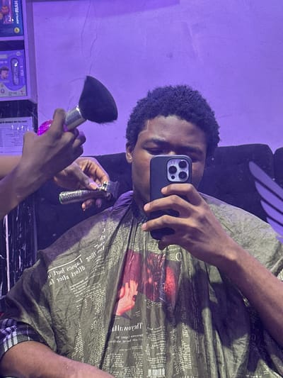 A person is taking a selfie in what appears to be a barber shop setting, as indicated by the bib around their neck, which is covered in newsprint and an image of hands. The person is holding a smartphone in one hand, likely taking the photo, while holding a duster brush and an electric hair trimmer with the other. The location appears to be indoors, possibly in Ogwashi-Uku, Nigeria, with a shelf of products visible at the bottom of the frame. The wall behind is painted a light shade of purple.