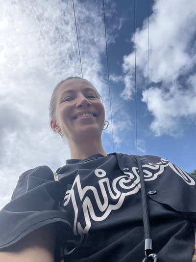 A woman is taking a selfie outdoors against a cloudy sky with power lines visible. She is wearing a black t-shirt with white lettering and large hoop earrings. She is smiling at the camera. The image does not contain any visual cues to suggest the location is Ahangama, Sri Lanka.