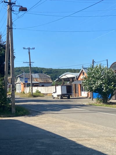 This is a daytime, eye-level, outdoor, medium shot of a street scene in Maykop, Russia. The scene features a paved road with white lane markings, bordered by a dirt shoulder and grass. A large power pole with multiple wires and a street lamp stands to the left. In the background, there are several buildings, including a house with a corrugated metal roof and brick accents, and another building with an orange facade. A white utility van with an open back is parked on the side of the road in front of these buildings. A flowering tree with white blossoms is prominent on the right side of the image. The sky is clear and blue. The overall impression is of a rural or suburban street on a sunny day.
