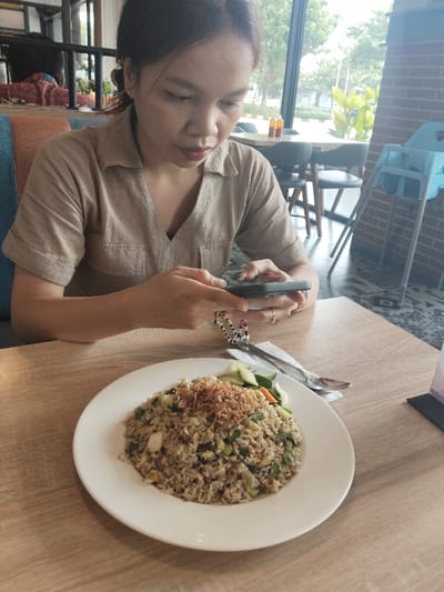 The image shows a woman in a light brown shirt sitting at a table in a restaurant, holding a smartphone in her right hand and looking at the screen. She is wearing a bracelet on her wrist. In front of her is a white plate with a serving of fried rice with vegetables, topped with fried onions. There is a napkin, two spoons, and a piece of cucumber and carrot on the plate. The table is made of light wood and there are additional tables and chairs behind the woman. The scene takes place in a restaurant in the city of Bekasi Regency, Indonesia, located at coordinates -6.299 latitude and 107.151 longitude. The restaurant features a large window overlooking a green area.