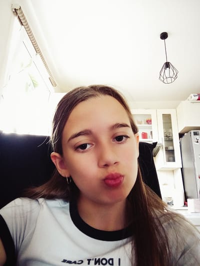 A young girl with long brown hair is taking a selfie in what appears to be a kitchen. She is wearing a white t-shirt with a black trim around the collar and a subtle inscription on the front. Her lips are pursed in a playful expression. The background shows white kitchen cabinets with glass fronts displaying some items, a refrigerator, and a window with blinds. A modern geometric pendant light hangs from the ceiling. The room is brightly lit, suggesting it's daytime. There are no visible landmarks that would specifically place the location in Radzymin, Poland.