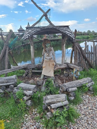 The image shows an outdoor scene featuring a rustic, whimsical structure built from wood and natural materials. A central figure, a doll or mannequin with green skin, long brown hair, and a somewhat sinister smile, is positioned under a thatched-roof shelter. The figure is adorned with necklaces made of shells and other organic materials and wears a tan, rough-textured skirt. The shelter is supported by rough-hewn wooden posts and beams, and the roof is made of overlapping wooden shingles.
The structure is situated near a body of water, likely a pond or lake, with tall reeds growing along its edge. A rustic wooden fence, made of vertically placed logs, borders the water. The foreground is filled with various logs and branches, some neatly stacked and others scattered, as well as lush green grass and small pebbles. In the background, the environment suggests a park or recreational area, with more rustic buildings and greenery visible. The sky above is partly cloudy with blue patches.
This scene is located in the village of Malakhovets, Malakhovetski Sel'ski Sovet, Belarus.