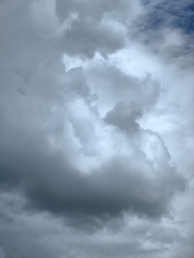A low-angle, full-frame shot captures a sky filled with a thick, overcast of grey clouds. The clouds are predominantly dark and heavy, with lighter, diffused areas showing through, suggesting the presence of sunlight behind them. There are no discernible human figures or man-made structures visible in the frame. The scene is natural and devoid of any urban elements, indicating it was taken in an open area with no reference points to a specific city location. The atmospheric conditions suggest a potentially rainy or stormy day.
