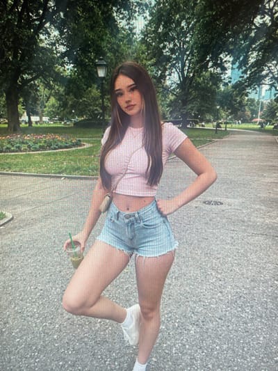 A young woman stands in a park, holding a drink. She has long, dark hair and is wearing a pink crop top and denim shorts. Her left hand is on her hip, and her right hand holds a clear plastic cup with a green straw. The park features lush green trees, a paved pathway, and flower beds in the background. A lamppost is visible among the trees. The scene suggests a casual outdoor setting. The location in the city of Mantov, Czechia, is not discernible from the provided image.