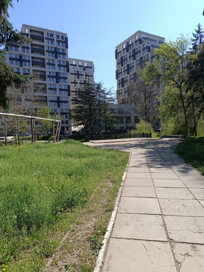 The image depicts an outdoor scene in Simferopol, Ukraine, featuring two high-rise buildings in the background. A concrete-tiled pathway extends from the foreground, leading towards a slightly elevated area with a metal railing. To the left of the pathway is a patch of green grass with some wildflowers, and further left, a metal structure resembling playground equipment or pipes is visible. Trees are scattered throughout the scene, adding greenery. The sky is clear and blue, suggesting a sunny day. No people are visible.