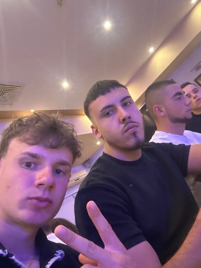 A selfie, taken at an upward angle, captures three young men indoors. The foreground figure has light skin, short brown hair, and is wearing a light-colored shirt. The middle figure has olive skin, short dark hair, a trimmed beard, and is wearing a black shirt. The background figure has olive skin, short dark hair, and is wearing a white shirt. The middle figure is making a duck-face expression. The foreground figure is making a peace sign with his fingers. The location is indoors, possibly a home or establishment in an unknown city. No specific evidence exists to establish that the photo was taken in the city of Elhovo, Bulgaria. The lighting is diffused and the background is partially obscured. A ceiling vent can be seen in the bottom left corner of the image.