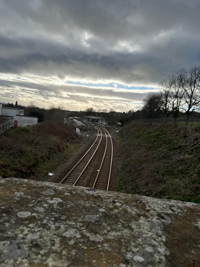 The image presents an elevated view of a railway track curving through a landscape. The tracks run diagonally from the upper-middle to the lower-middle of the frame. To the left of the tracks, buildings and parked cars are visible. Overlooking the tracks, there is a slope covered with vegetation and trees. The sky, displaying cloud formations, is situated on the left-hand side. In the foreground on the right is an out-of-focus, lichen-covered wall or stone structure, likely a part of a bridge or similar elevated construction. The scene is in Dunfermline, United Kingdom.