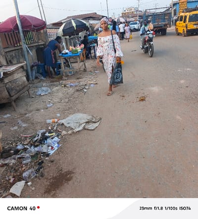 The image captures an outdoor street scene in Agbani, Nigeria, likely during late afternoon or early evening. In the foreground, a young woman walks along a dirt and paved road, slightly smiling at the camera. She wears a white, off-shoulder dress with a floral pattern and a matching headwrap, holding a black plastic bag with a prominent "S" logo. Her feet are visible in sandals.
To her left, a market stall is set up under a dark umbrella, with a woman in a blue dress attending to fruits and vegetables, including watermelons and cucumbers, displayed on a wooden table. Another red umbrella is visible further back. The ground near the market stalls and along the roadside is littered with debris, plastic waste, and dirt.
In the mid-ground and background, the street continues with ongoing activity. A man on a motorcycle drives past the main subject, and other pedestrians are visible further down the road. Several vehicles populate the street, including a yellow minibus (characteristic of public transport in Nigeria) and a blue truck. Buildings line the street, suggesting a commercial or residential area. The lighting is soft, indicating either early morning or late afternoon.
Visible text includes "CAMON 40" and "23mm f/1.8 1/100s ISO74" at the bottom of the image, detailing the camera model and settings. The black bag held by the main subject also shows an "S" logo.