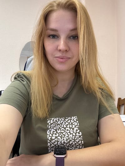 A young woman with shoulder-length blonde hair and fair skin is seated at a desk, looking directly at the camera. She is wearing a green t-shirt with a white, abstract pattern. Her left arm is crossed over her right arm, and she is wearing a purple watch on her left wrist. In the background, a wall with a textured finish is visible, along with a round mirror reflecting a portion of the room. To the right, a wooden chair is partially visible. The lighting suggests an indoor setting, possibly an office or a home study. The image does not provide any specific indicators of its location within the city of Заволжье, Russia, beyond the fact that it was taken there.