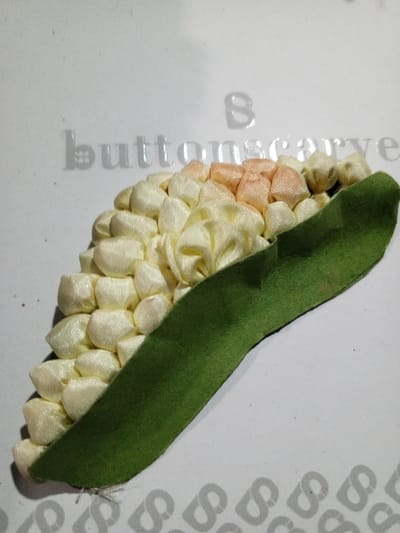 A ribbon-crafted ear of corn rests on a white surface with the faint imprint of "buttonscarve" visible. The corn features a cream-colored kernel base, transitioning to peach-colored kernels at the top. A bright green husk curves along the side, adding a naturalistic contrast. The individual kernels are tightly packed, displaying a delicate, handmade quality. The overall composition presents a charming, artistic rendering of the vegetable.