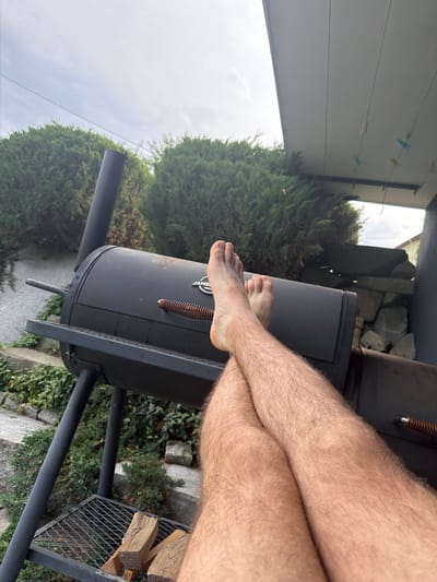 A person's bare legs and feet are crossed over a black barbecue smoker in a backyard setting. The smoker has a side stack and a front handle. Wood logs are stacked on a metal rack beneath the smoker. In the background, there are green bushes, stone landscaping, and a partial view of a building. The sky is overcast. The image is taken from a low-angle perspective, focusing on the legs and the smoker.