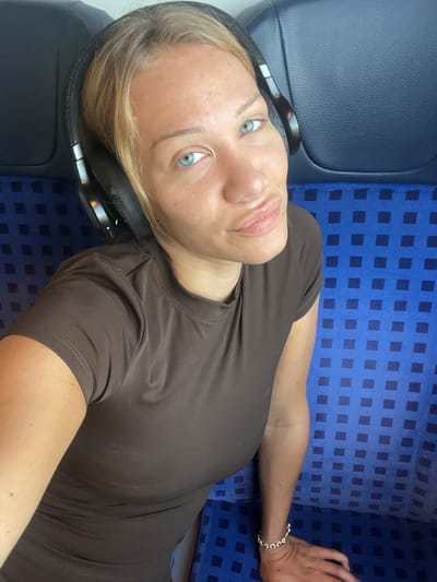 A woman with blonde hair, blue eyes, and headphones over her ears is taking a selfie on a train. She is wearing a brown t-shirt and a bracelet on her left wrist. The train seats are upholstered in blue fabric with a dark blue checkered pattern. The image is taken from a high angle, looking down at the woman. It is unclear where in Ampfing, Germany this photo was taken, but it appears to be inside a public transportation vehicle.