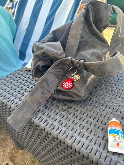 A gray leather shoulder bag sits on a woven patio chair. The bag features a thick strap with a silver buckle and a red mushroom-shaped keychain. A tube of sunscreen rests on the chair next to the bag. Part of a blue and white striped cloth and light blue fabric are visible in the lower left corner. The chair is dark gray with a woven texture.