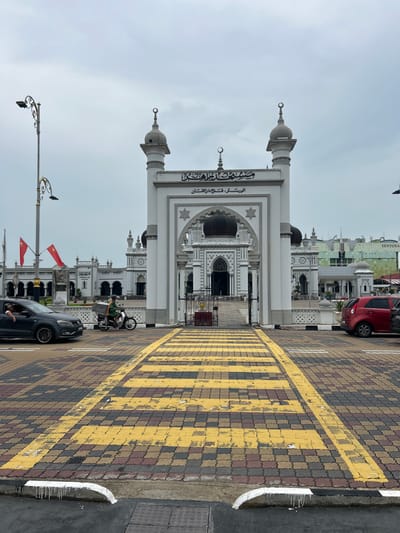 The image depicts the Zahir Mosque in Alor Setar, Malaysia, on an overcast day. The mosque features white architecture with dark domes and minarets, adorned with Islamic calligraphy above its main entrance archway. In the foreground, a pedestrian crossing with yellow and black stripes is laid out on a patterned brick surface. Two cars, one dark grey and one red, are parked on the right side of the frame, partially obscuring the view of the mosque's side. A man in a green shirt is riding a motorcycle with a storage box on the back, crossing the street from left to right. Another man is visible in the driver's seat of the dark grey car on the left. Red flags with a crescent moon and star emblem are planted on poles to the left of the mosque's entrance. In the background, beyond the mosque, a modern building with the "SENTOSA" logo is visible. The overall scene captures a moment of everyday life in front of a significant religious landmark.
