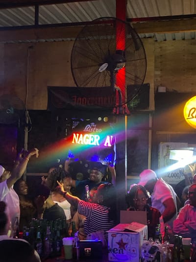 The scene is set in a dimly lit bar in Dodoma, Tanzania. The bar is decorated with a large, black ceiling fan and a sign advertising "The Bistro Nager A." There is a rainbow effect emanating from the sign, creating a colorful visual. The bar is bustling with a group of people, likely friends or patrons, interacting and socializing. A man wearing a white shirt stands at the bar. A woman in a striped shirt takes a selfie with her phone while engaging with other patrons. Several other people are seated, and there are numerous bottles of beer and other alcoholic beverages on the counter. A person behind the bar is obscured by a large box of beer, but their arm and hands are visible. The environment is lively and vibrant, with various people engaged in conversation and enjoyment. The bar has an open-air design, with a corrugated metal roof. The lighting is primarily artificial, with dim lights casting shadows around the room, and the back wall is covered in brown paneling. The bar is an informal gathering place where patrons can socialize and enjoy refreshments.