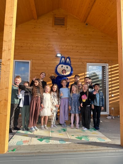 In Oseyevo, Russia, a group of children and an adult in a blue mascot costume are gathered on the wooden porch of a house. The mascot, which appears to be a blue cat or similar creature with large eyes and ears, stands in the center holding a cake with a single candle. The children, ranging in age, are positioned around the mascot, some smiling, some with mouths open in exclamations, and one giving a peace sign. The house has a wooden exterior and a pitched roof with a vent. Sunlight casts shadows across the porch, which is covered by a mat with a printed pattern of shapes and figures. A few pairs of shoes are visible on the edge of the porch.