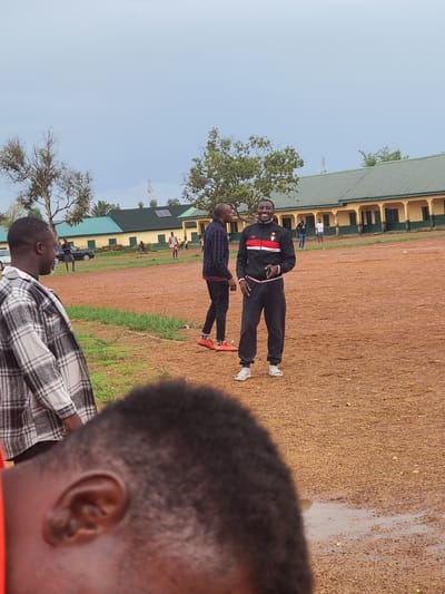 This daytime image, captured outdoors in Rafin Bauna, Nigeria, features a scene on a reddish-brown dirt and gravel field. In the immediate foreground, the right side of a person's head and ear is visible, with short dark hair, and they are wearing a black and white plaid shirt, seemingly observing the activity.
In the mid-ground, two men stand on the field. The man closer to the viewer is dressed in a dark, possibly plaid, long-sleeved shirt, dark pants, and red and black sneakers. He faces the central figure, a smiling man wearing a black tracksuit with prominent red and white stripes and a star-like emblem on the chest. This man is wearing white sneakers and appears to be looking towards the camera.
The background is dominated by a long, two-story building with a green corrugated metal roof and light-colored walls, featuring numerous windows or doorways. Several other individuals are scattered across the field and near the buildings, including one person in a white shirt and dark pants. A dark-colored car is partially visible near the structures. Trees with varying degrees of foliage are present across the landscape. A distinct wet patch or puddle is visible on the dirt field, indicating recent rain or standing water. A communication tower can be seen on the distant horizon against a clear, bright sky. The scene depicts a casual outdoor gathering or meeting. No text is visible in the image.