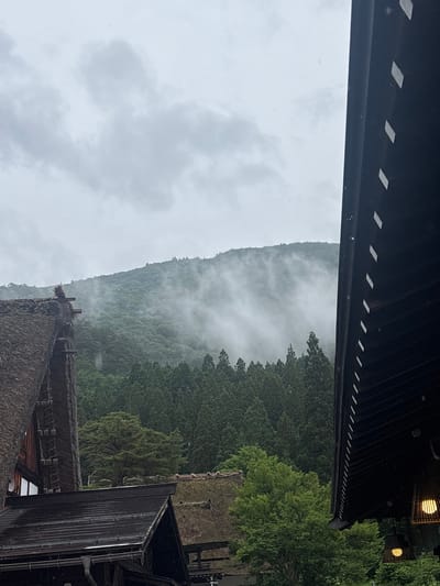 This vertical image captures a misty, overcast day in Shirakawa, Japan, showcasing traditional architecture against a mountainous backdrop. The upper portion of the frame is filled with a pale grey, featureless sky, indicating heavy cloud cover. Below this, a large, densely forested mountain range rises, with its mid-section significantly obscured by thick white mist or fog. The visible lower slopes and mid-ground are covered in various green trees, including numerous dark green conifers and lighter green deciduous trees.

In the left foreground, a prominent section of a traditional gassho-zukuri house is visible. Its steeply pitched roof is heavily thatched with dark, textured straw, showing some wear and exposed wooden structural elements near the peak. In the right foreground, the dark, multi-layered eaves and part of the side of another traditional building are seen, featuring a dark, possibly tiled or planked roof. Along the underside of these eaves, a series of evenly spaced, horizontal light-colored rectangular elements are present. Numerous small, reflective droplets, consistent with rain or heavy mist, are visible adhering to and dripping from these eaves. Below the eaves, two illuminated, lantern-style light fixtures, constructed from a woven material resembling bamboo, hang and emit a warm, yellow glow. Parts of additional traditional roofs, some thatched and others with dark, corrugated or tiled surfaces, are visible in the lower-mid ground, nestled amongst the trees. No human figures are present.