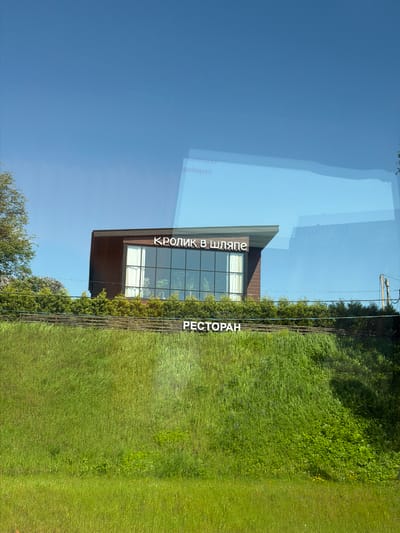 A photograph shows the building housing the restaurant "Krolik v Shliape" (Кролик в Шляпе) in Yanino-1, Russia. The building is a modern structure with dark wood siding and a large glass window façade. The restaurant's name is displayed prominently above the window. A green hedge and tall grass grow along the front of the building. Blue sky is visible in the background. Overhead wires are also visible. The image is shot from a slightly low angle, and there are reflections in the glass. The image appears to be taken from inside a vehicle, as there is a reflection on the glass.