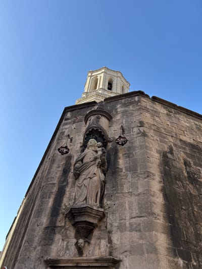 This is a low-angle, medium shot of the exterior of a stone building, likely a church, in Girona, Spain. The building's corner is the main subject, with rough-hewn grey stone blocks forming its facade. A weathered stone statue of the Virgin Mary holding the infant Jesus is mounted in a niche on the corner. The statue is draped in flowing robes and has a serene expression. Below the statue, a small, winged cherub head carving is visible. Two antique, wrought-iron hanging lamps with intricate designs are attached to the wall on either side of the niche. Above the corner of the building, a portion of a white stone bell tower with arched openings and a rounded top is visible against a clear, bright blue sky. The stone of the building shows signs of age and weathering, with dark streaks staining the lower sections of the facade. A hint of a person, out of focus, can be seen in the lower left corner of the frame. The overall impression is one of historical architecture and religious art.