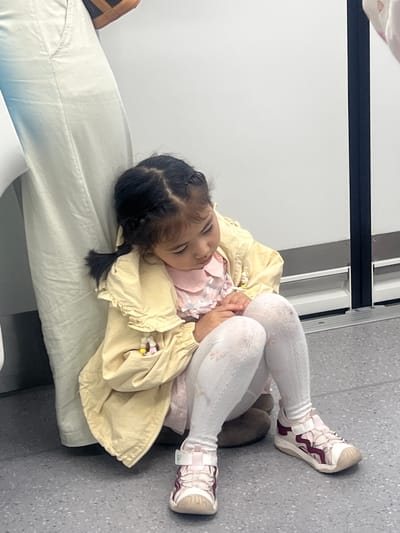 A young girl with dark hair in braids is sleeping on a white cushioned bench in an indoor setting, likely a waiting area. She wears white patterned leggings, pink and white sneakers, a pink shirt, and a yellow jacket. Her legs are crossed, and her hands are clasped near her chest. The bench is set against a gray wall and floor. Overhead, there is a black bar or rail. The image does not provide enough information to determine that the location is the city of Tiaodeng, China.