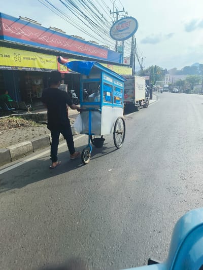 The scene is a street in Purwakarta, Indonesia, with coordinates of -6.561 latitude and 107.437 longitude. A man is walking on the street, pushing a food cart. The cart is blue and white, with a sign that reads "Sosis Bakso". The cart has two wheels and a handle. The man is wearing a black shirt and blue jeans. He is walking away from the camera towards a white van on the side of the street. The street is paved with asphalt and lined with buildings on both sides. The sky is clear and sunny. In the background there is a sign for "Planet Ban". There are several cars parked on the side of the street, one of which is a white van. There is a small green area on the left side of the street, just beyond the curb. There are some green hills in the distance.