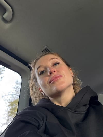The image shows a woman with light skin, curly blonde hair, and defined eyelashes. She is wearing a black hooded sweatshirt. The perspective is from below, looking up at her face. The background includes the interior of a vehicle, specifically the ceiling and part of a window. Outside the window, blurred trees are visible, suggesting an outdoor environment. There is no evidence to suggest this is in Achadara, Abkhazia.